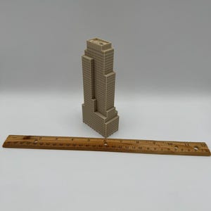 May include: A beige miniature skyscraper model with a ruler. The building has a rectangular shape with multiple levels and a flat roof. The wooden ruler is marked with inch measurements.