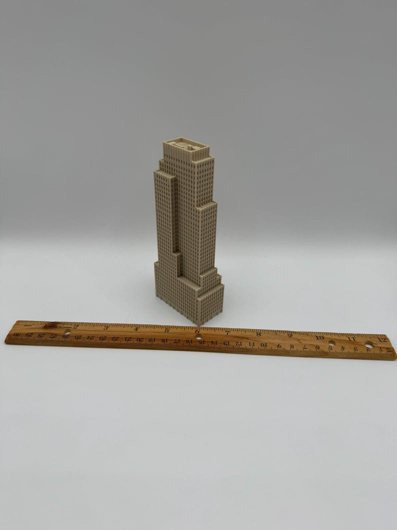 May include: A beige miniature skyscraper model with a ruler. The building has a rectangular shape with multiple levels and a flat roof. The wooden ruler is marked with inch measurements.