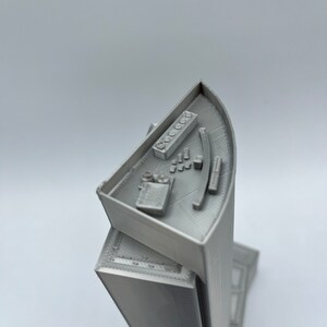 May include: A 3D printed model of a skyscraper with a detailed rooftop and a curved facade. The rooftop features a small control panel with buttons and a curved railing.