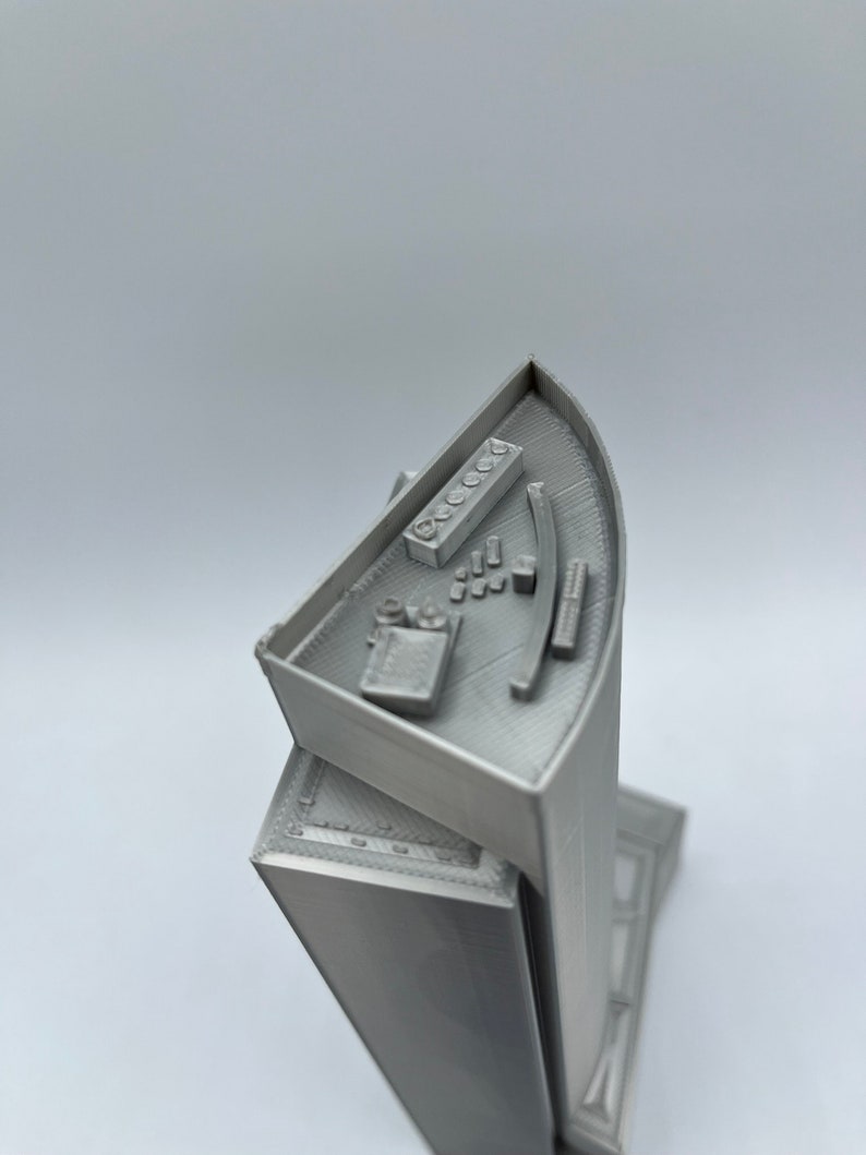 May include: A 3D printed model of a skyscraper with a detailed rooftop and a curved facade. The rooftop features a small control panel with buttons and a curved railing.