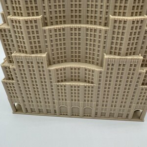 May include: A beige architectural model of a multi-tiered building, featuring numerous windows and a detailed facade. The design includes arched entryways and a stepped structure, resembling a classic skyscraper.