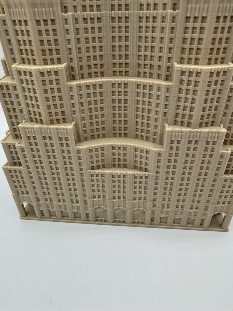 May include: A beige architectural model of a multi-tiered building, featuring numerous windows and a detailed facade. The design includes arched entryways and a stepped structure, resembling a classic skyscraper.