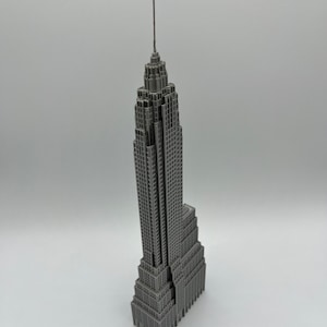 K&ouml;nnte beinhalten: Ein graues Miniaturmodell des Empire State Building. Das Modell weist detaillierte architektonische Elemente auf, darunter die Antenne und mehrere Ebenen. Das Geb&auml;ude hat eine einheitliche graue Farbe und steht vor einem wei&szlig;en Hintergrund.