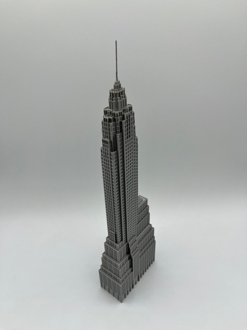 K&ouml;nnte beinhalten: Ein graues Miniaturmodell des Empire State Building. Das Modell weist detaillierte architektonische Elemente auf, darunter die Antenne und mehrere Ebenen. Das Geb&auml;ude hat eine einheitliche graue Farbe und steht vor einem wei&szlig;en Hintergrund.