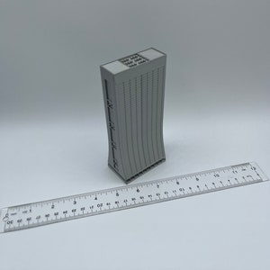 May include: A gray 3D printed model of a tall building with a curved facade and a flat roof. The model is approximately 6 inches tall and is sitting on a white surface.