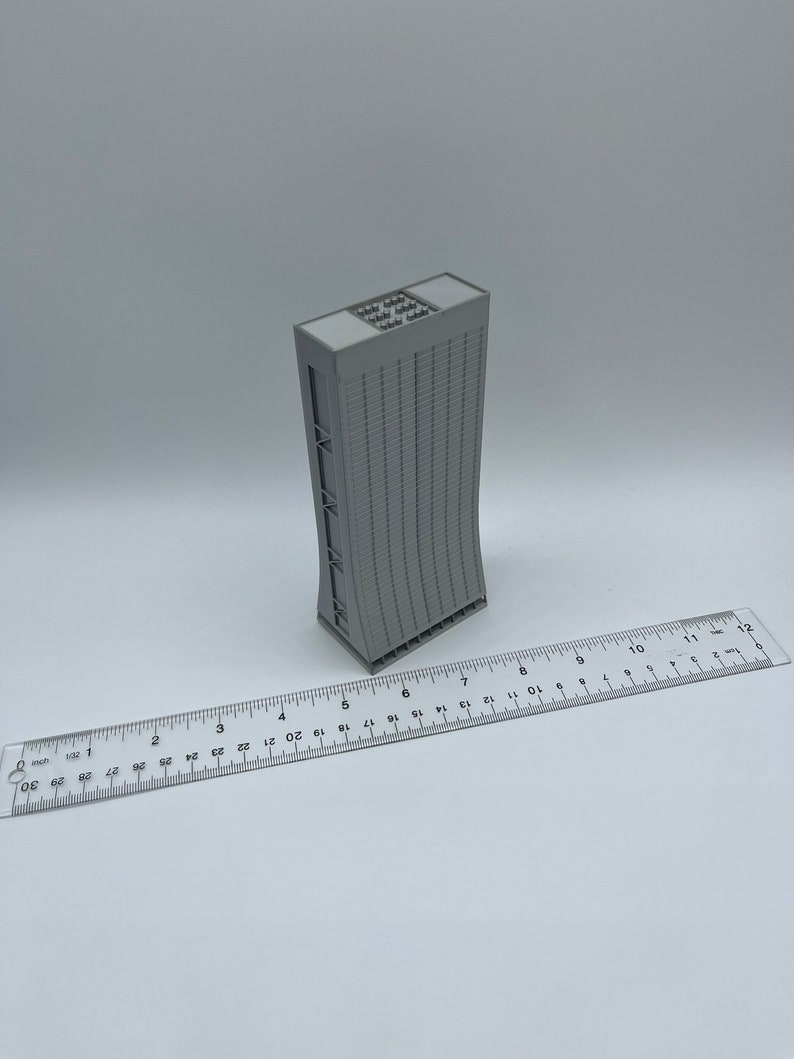 May include: A gray 3D printed model of a tall building with a curved facade and a flat roof. The model is approximately 6 inches tall and is sitting on a white surface.