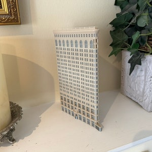 Op de afbeelding: Een wit miniatuurmodel van een hoog gebouw met veel ramen. Het model staat op een wit rek met een witte kaars en een potplant op de achtergrond.