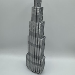 May include: A silver-colored 3D printed model of a skyscraper featuring a stepped, tiered architectural design. The structure displays vertical lines, rectangular window patterns, and prominent diagonal cross-bracing details on the exterior facade.