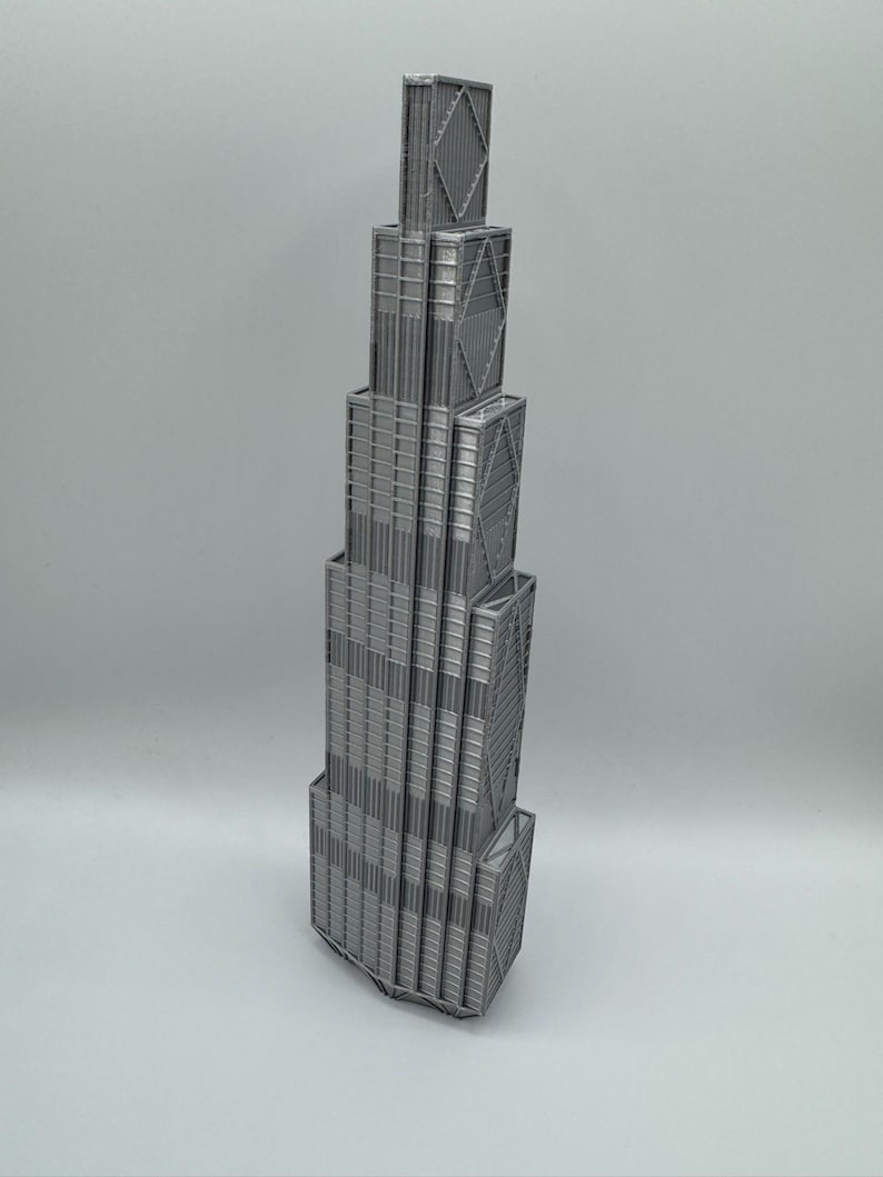 May include: A silver-colored 3D printed model of a skyscraper featuring a stepped, tiered architectural design. The structure displays vertical lines, rectangular window patterns, and prominent diagonal cross-bracing details on the exterior facade.