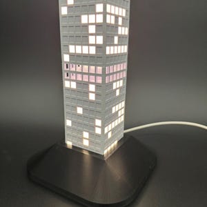 May include: A gray, rectangular lamp shaped like a skyscraper, with illuminated windows. The lamp sits on a black base and has a white cord. The windows are lit with a warm, inviting glow, creating a modern, architectural design.