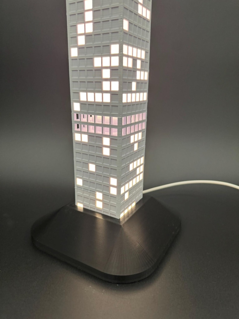 May include: A gray, rectangular lamp shaped like a skyscraper, with illuminated windows. The lamp sits on a black base and has a white cord. The windows are lit with a warm, inviting glow, creating a modern, architectural design.