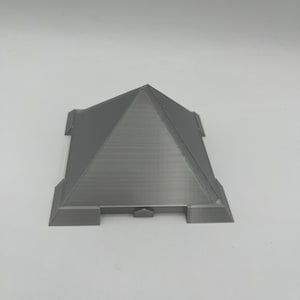 Puede incluir: Un objeto gris en forma de pir&aacute;mide impreso en 3D, con una base plana y una parte superior puntiaguda. La base tiene cuatro extensiones rectangulares. La pir&aacute;mide tiene l&iacute;neas horizontales visibles del proceso de impresi&oacute;n. El objeto est&aacute; sobre un fondo blanco.