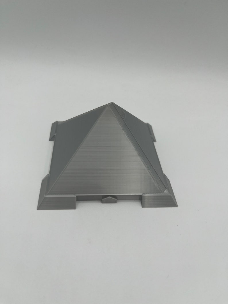 Puede incluir: Un objeto gris en forma de pir&aacute;mide impreso en 3D, con una base plana y una parte superior puntiaguda. La base tiene cuatro extensiones rectangulares. La pir&aacute;mide tiene l&iacute;neas horizontales visibles del proceso de impresi&oacute;n. El objeto est&aacute; sobre un fondo blanco.