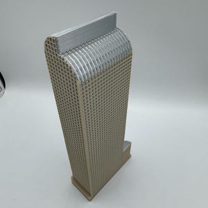 May include: A beige, 3D-printed architectural model of a skyscraper. The building has a rectangular base and a tall, grid-like structure. The top of the building has a curved design with a silver-colored accent.