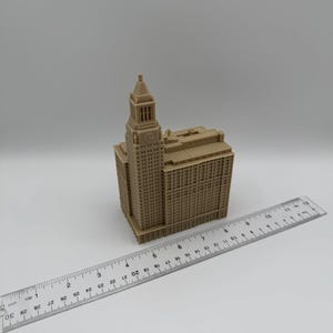 May include: A tan-colored miniature building with a clock tower, resembling a skyscraper. A ruler is placed next to the building, measuring approximately 3 inches. The model has detailed architectural features.