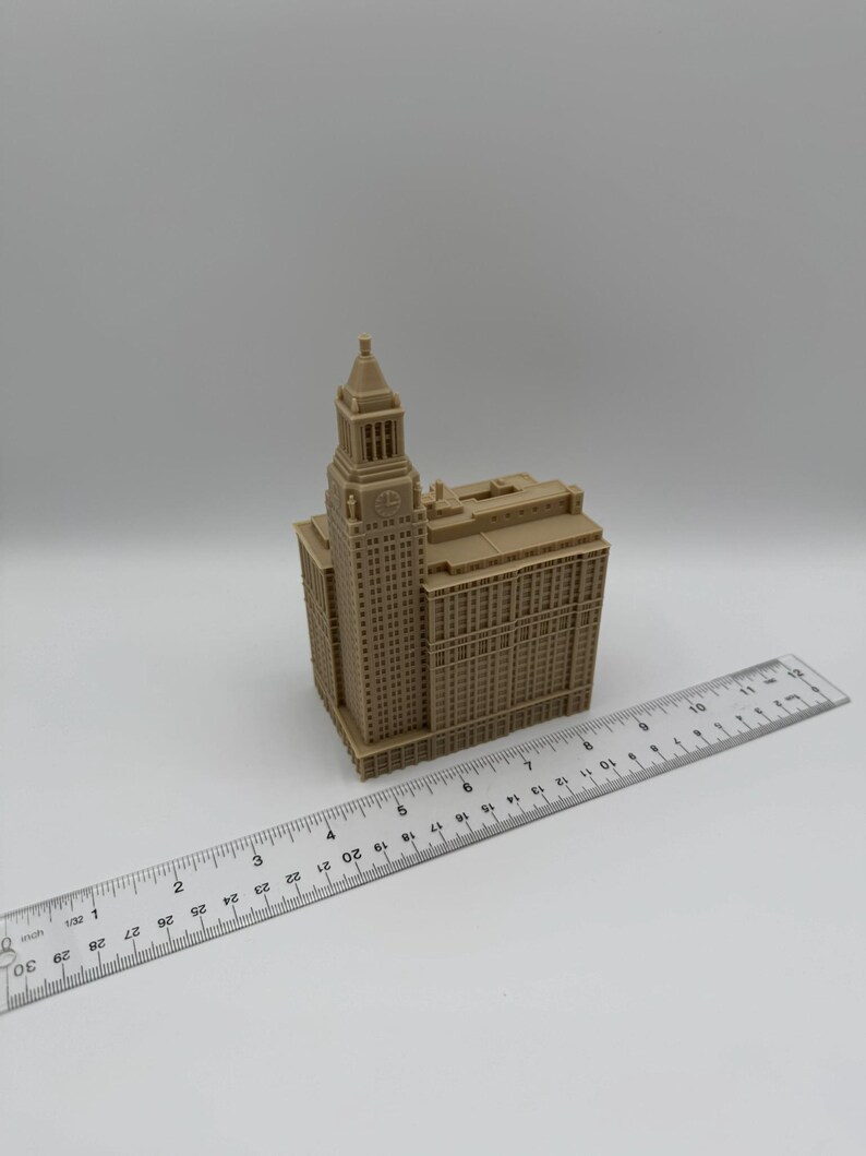 May include: A tan-colored miniature building with a clock tower, resembling a skyscraper. A ruler is placed next to the building, measuring approximately 3 inches. The model has detailed architectural features.