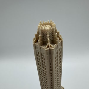 May include: A beige architectural model of a tall building with many windows. The model has a detailed top with intricate designs and a rectangular base. The building's facade is textured, and the overall design is symmetrical.