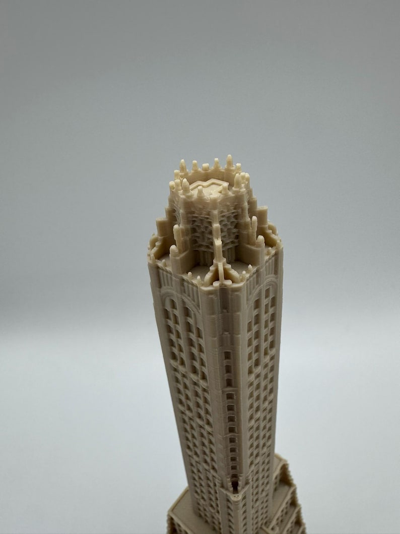 May include: A beige architectural model of a tall building with many windows. The model has a detailed top with intricate designs and a rectangular base. The building's facade is textured, and the overall design is symmetrical.