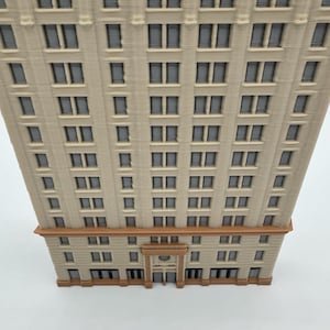 May include: A detailed model of a multi-story building with numerous windows. The building has a beige facade with brown accents at the base and top. The windows are dark, and the overall design suggests a classic architectural style.