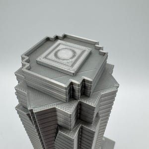 May include: A gray, 3D-printed model of a skyscraper. The building has a layered design with a square top and a series of stepped levels. The top features a recessed square with a circular detail. The model is made of a solid, light gray material.