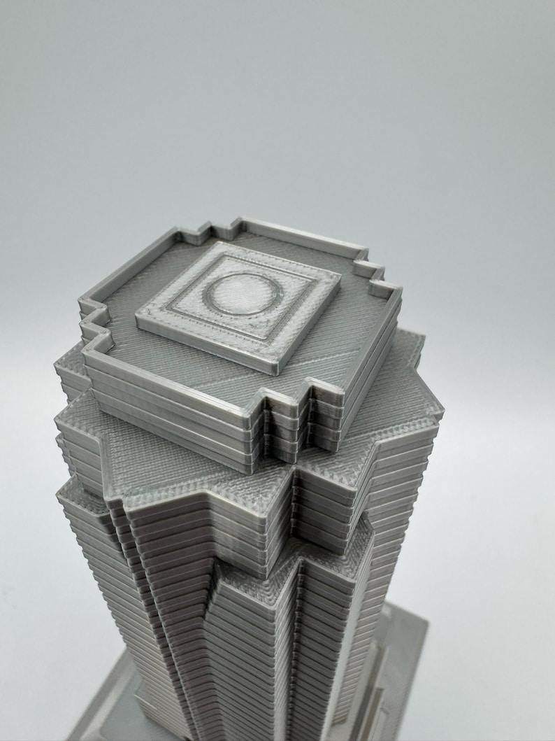 May include: A gray, 3D-printed model of a skyscraper. The building has a layered design with a square top and a series of stepped levels. The top features a recessed square with a circular detail. The model is made of a solid, light gray material.