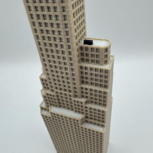 May include: A beige 3D printed model of a skyscraper featuring a stepped architectural design with multiple levels, numerous rectangular window cutouts, and a small black rectangular block positioned on the flat roof of the middle section.