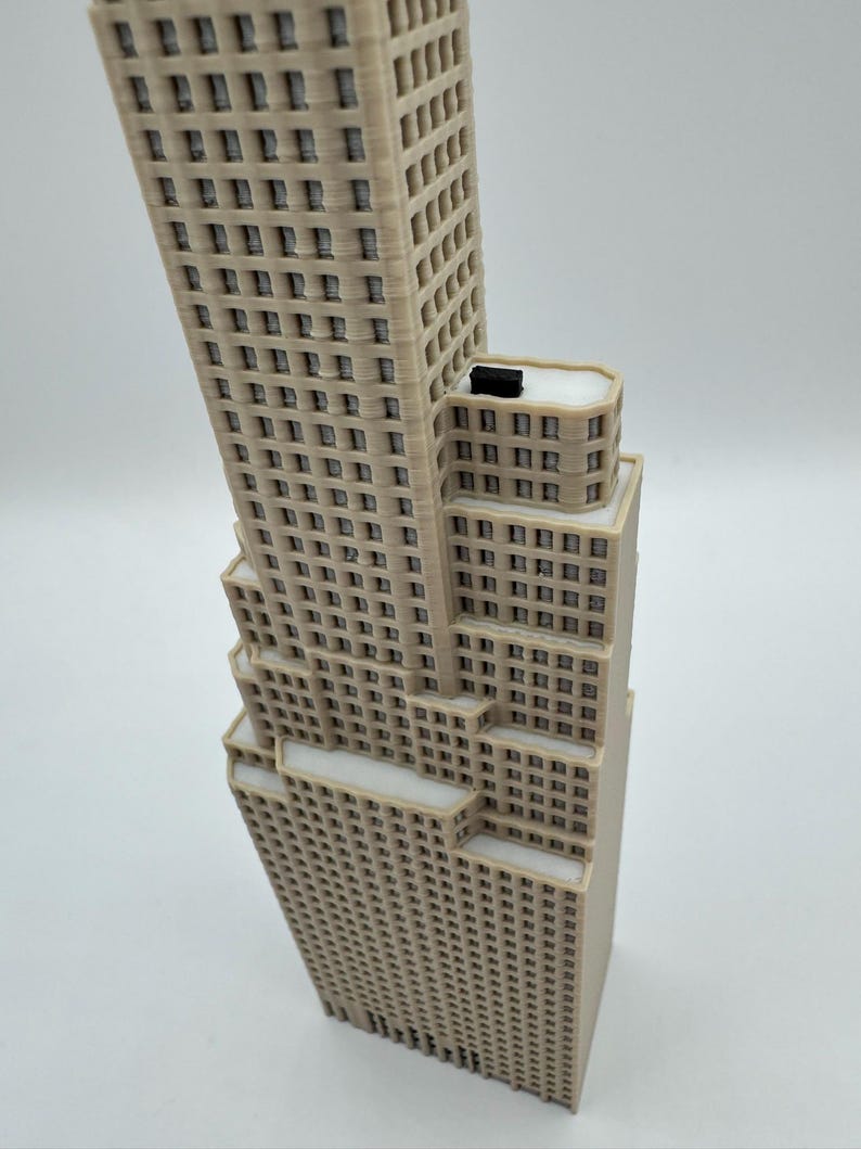May include: A beige 3D printed model of a skyscraper featuring a stepped architectural design with multiple levels, numerous rectangular window cutouts, and a small black rectangular block positioned on the flat roof of the middle section.