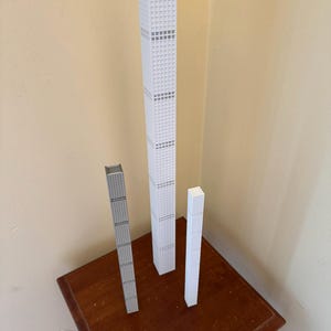 May include: Three miniature skyscraper models in white and grey, standing on a dark wooden table. The tallest model is white with grey accents, while the other two are grey and white. A clear ruler lies in front.