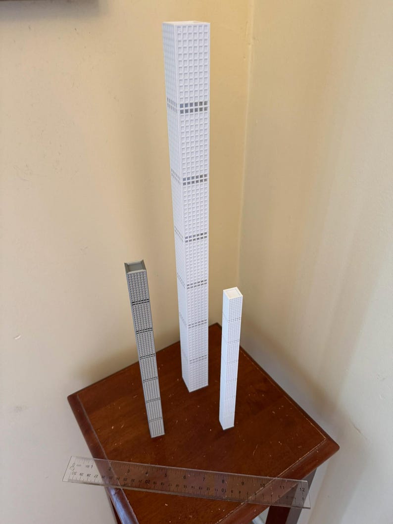 May include: Three miniature skyscraper models in white and grey, standing on a dark wooden table. The tallest model is white with grey accents, while the other two are grey and white. A clear ruler lies in front.