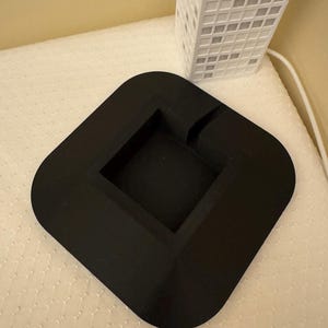 May include: A black, square ashtray with rounded corners and a recessed center. A white, rectangular building with a grid pattern is in the background. The ashtray is on a white, textured surface.
