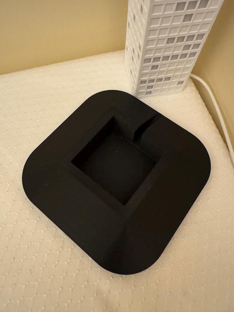 May include: A black, square ashtray with rounded corners and a recessed center. A white, rectangular building with a grid pattern is in the background. The ashtray is on a white, textured surface.