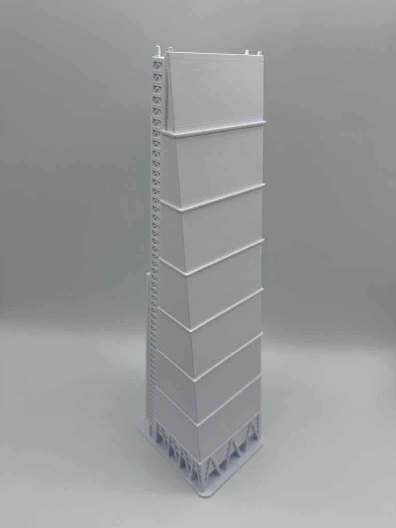 May include: A tall, white, architectural model of a grain silo or industrial structure. The model has a rectangular shape with multiple stacked sections and a ladder running along one side. The base is a triangular shape.