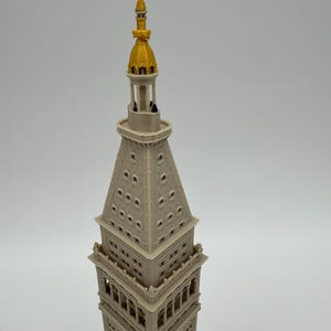 May include: A 3D printed scale model of the Metropolitan Life Insurance Company Tower. The structure is light beige with a prominent gold-colored spire at the top. It features detailed architectural elements including windows and clock faces.
