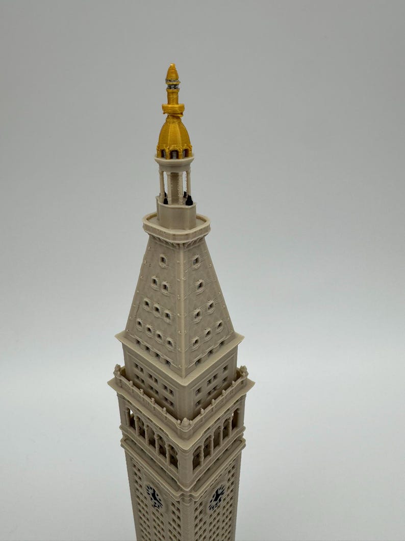 May include: A 3D printed scale model of the Metropolitan Life Insurance Company Tower. The structure is light beige with a prominent gold-colored spire at the top. It features detailed architectural elements including windows and clock faces.