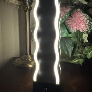 K&ouml;nnte beinhalten: Eine schwarze, rechteckige LED-Lampe mit einem wellenf&ouml;rmigen, beleuchteten wei&szlig;en Design. Die Lampe steht auf einer dunklen Oberfl&auml;che, mit einem Kabel, das von der Basis ausgeht. Eine Vintage-Lampe und Gr&uuml;nzeug sind im Hintergrund.