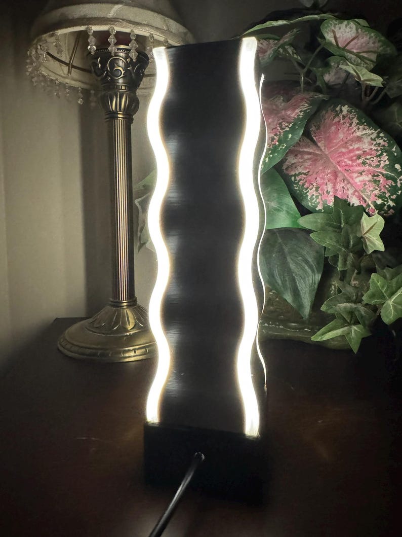K&ouml;nnte beinhalten: Eine schwarze, rechteckige LED-Lampe mit einem wellenf&ouml;rmigen, beleuchteten wei&szlig;en Design. Die Lampe steht auf einer dunklen Oberfl&auml;che, mit einem Kabel, das von der Basis ausgeht. Eine Vintage-Lampe und Gr&uuml;nzeug sind im Hintergrund.