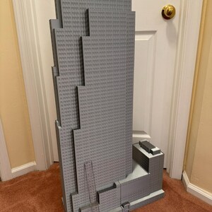 May include: A gray model of a skyscraper with a detailed facade, featuring numerous windows and architectural details. The model is a light gray color and stands tall, showcasing the building's design. The model is placed on a brown carpet.