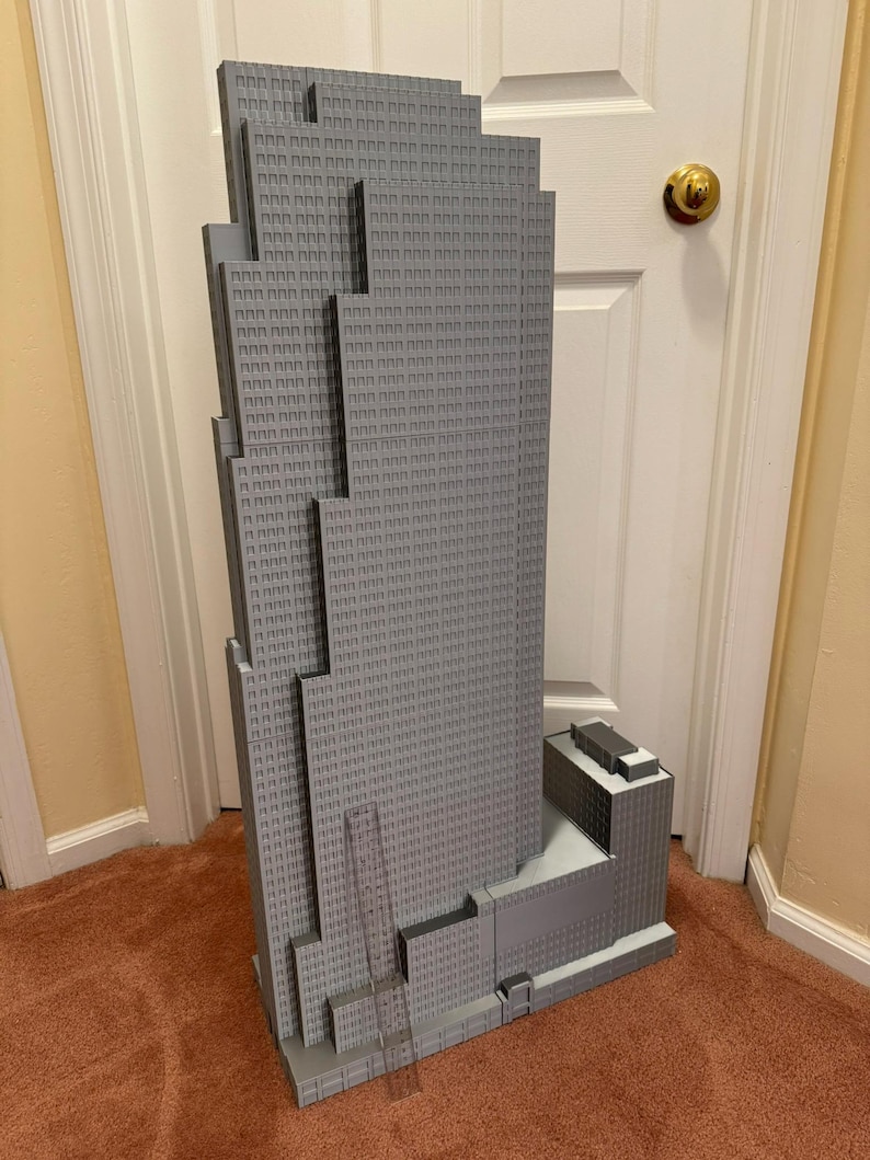 May include: A gray model of a skyscraper with a detailed facade, featuring numerous windows and architectural details. The model is a light gray color and stands tall, showcasing the building's design. The model is placed on a brown carpet.