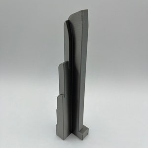 May include: A gray, 3D-printed architectural model with a layered design. The model features a rectangular base and a series of vertical, curved elements. The overall structure is approximately 6 inches tall.