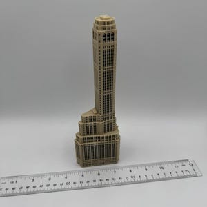 Op de afbeelding: Een beige miniatuurmodel van een hoge wolkenkrabber met een gedetailleerde gevel. Het model is ongeveer 15 cm hoog en staat op een liniaal.
