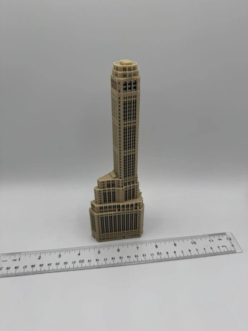 Op de afbeelding: Een beige miniatuurmodel van een hoge wolkenkrabber met een gedetailleerde gevel. Het model is ongeveer 15 cm hoog en staat op een liniaal.