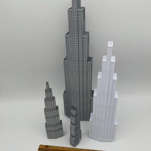 May include: Four 3D-printed skyscraper models in gray and white, featuring tiered architectural designs with vertical lines. A wooden ruler is placed in front, showing the tallest model is approximately 12 inches high and the smallest is 4 inches.