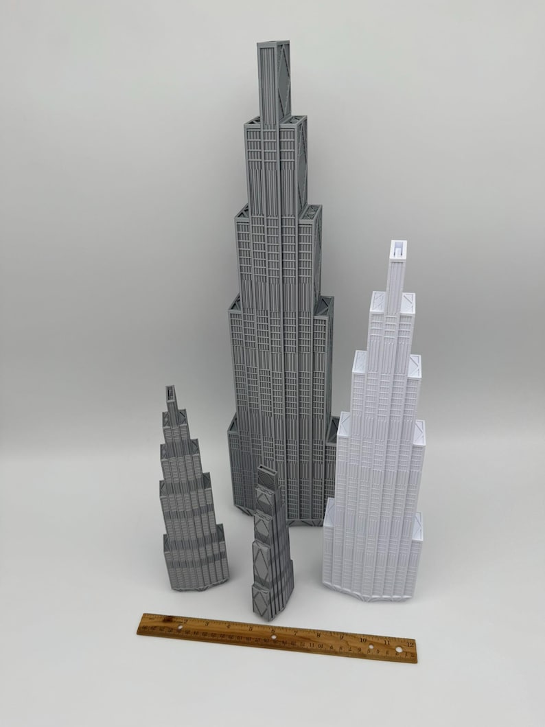May include: Four 3D-printed skyscraper models in gray and white, featuring tiered architectural designs with vertical lines. A wooden ruler is placed in front, showing the tallest model is approximately 12 inches high and the smallest is 4 inches.
