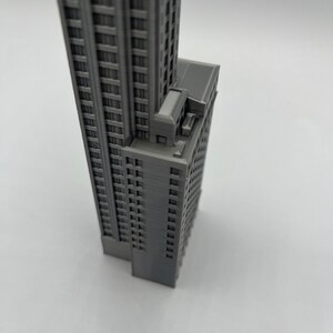 May include: A gray 3D printed model of a tall building with many windows. The building has a flat roof and a small, rectangular structure on the side.