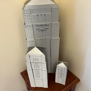 Peut inclure: Trois mod&egrave;les de gratte-ciel imprim&eacute;s en 3D de couleur grise. Le b&acirc;timent le plus haut porte l'inscription "WAYNE" sur le c&ocirc;t&eacute;. Les b&acirc;timents sont pos&eacute;s sur une surface en bois avec une r&egrave;gle.