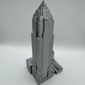 May include: A gray, 3D-printed model of a skyscraper. The building has a tiered design with a pointed top and detailed architectural features. The model is set against a plain white background.