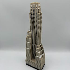 May include: A detailed miniature skyscraper model in shades of beige and off-white, with a black base. The building features numerous windows and architectural details, showcasing a classic design. The model stands tall against a plain background.