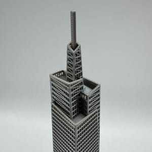 May include: A gray, 3D-printed model of a skyscraper, featuring a tall, rectangular main structure with multiple tiers and a prominent antenna. The building has a lattice-like design, showcasing intricate details.