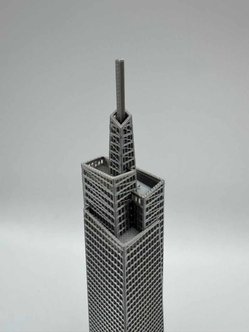 May include: A gray, 3D-printed model of a skyscraper, featuring a tall, rectangular main structure with multiple tiers and a prominent antenna. The building has a lattice-like design, showcasing intricate details.