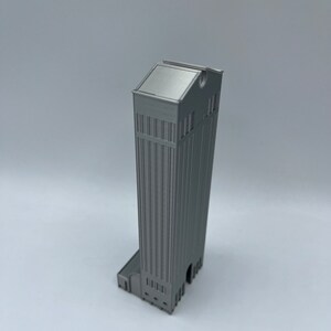 May include: A silver 3D printed model of a tall building with a flat roof and a rounded top. The building has a detailed facade with windows and a small entrance.